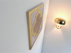 miroir décoratif carré beige avec cadre doré effet matelassé accroché sur mur blanc à côté d'applique dorée